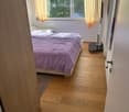 Bedroom with wooden flooring, a bed with purple bedding, a small table with a screen, and orange curtains on the windows.
