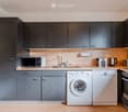 Modern kitchen with grey cabinets, white appliances including a microwave, washing machine, dishwasher, and oven, and a light wooden countertop.