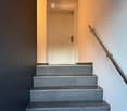 Staircase with grey tiled steps, white walls, and a metal handrail leading to a closed door at the top.