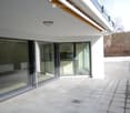 Modern balcony with large glass sliding doors leading to a kitchen area, tiled floor, and a scenic view of a wooded area.