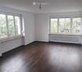 Empty room with wooden flooring, white walls, large windows, radiator, and a hanging light fixture.