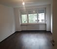 Empty room with wooden flooring, large window with blinds, radiator, and a hanging light fixture.