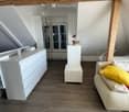 Bright, modern living room with wooden floors, white walls, and exposed wooden beams. Features a white sofa with yellow cushions, a small white cabinet, a stool, and a flower pot on a table.