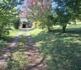 A gravel pathway leads to a small white house surrounded by lush greenery and trees, with a grassy lawn and a small porch area visible.