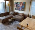 Modern living room with large windows offering a scenic view, featuring a brown sectional sofa, two small round tables, a wooden dining table with chairs, a floor lamp, and a colorful painting on the wall.