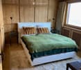 Cozy bedroom with wooden walls and ceiling, a large bed with green and white bedding, nightstands with lamps, and a fluffy white rug on the floor.