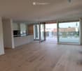 Open-plan unfurnished room with light wooden flooring, white walls, and large glass sliding doors leading to a balcony. A small kitchen area is visible with white cabinets and a countertop.
