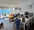 Bright living room with a view, featuring a sofa, dining table, chairs, vase with flowers, handbag, picture frame, and floor lamp.