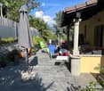 A well-maintained patio area with stone tiles, outdoor furniture including chairs and a table, potted plants, and an umbrella. The area is surrounded by greenery and has a view of mountains in the background.