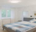 Modern bedroom with a large bed, nightstand, and mirror. The bed features a blue and white checkered duvet and matching pillows. The room has large windows with white curtains and a sleek, contemporary design.