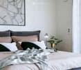 Modern bedroom with a large bed, multiple pillows, a nightstand with a lamp, a small plant, and a decorative picture frame on the wall.