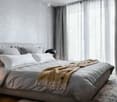 Modern bedroom with a large bed, multiple pillows, a blanket, and a nightstand with a lamp. The room features large windows with curtains and a geometric patterned wall.