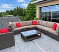 Outdoor patio with a modern wicker sectional sofa, red throw pillows, and a glass coffee table, overlooking a lush green landscape.