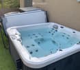 A modern hot tub with multiple jets and a control panel, situated on a grassy area next to a house with beige walls and a covered electrical unit.