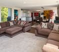 Modern living room with large windows offering a view of greenery, featuring a sectional sofa with pillows, an ottoman, a dining area with a table and chairs, and decorative art pieces.