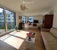 Bright living room with wooden flooring, large windows, and a balcony. Features a sofa, coffee table, armchair, ottoman, floor lamp, table lamp, television, cabinet, vase with flowers, and plants.