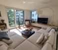 Modern living room with large windows and balcony access, featuring a grey sectional sofa, coffee table, armchair, TV stand, floor lamp, and decorative items.