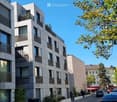 Modern multi-story apartment building with balconies, large windows, and contemporary design, situated on a street with trees and parked cars.