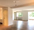 Empty room with wooden flooring, white walls, and large windows allowing natural light. Features include a radiator, a small cabinet, and a pendant light fixture.