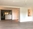 Empty room with light wooden flooring and white walls, featuring a modern pendant light fixture and an open doorway leading to a kitchen area.