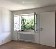 Empty room with light-colored walls, wooden flooring, a radiator, and a large window with a view of greenery and a neighboring house.