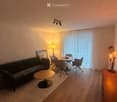 Modern living room with a grey sofa, coffee table, dining table with chairs, and decorative lamps. Large window with sheer curtains and a view of the balcony.