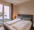 Modern bedroom with a large bed, headboard, pillows, and a nightstand near a large window with a view of a balcony area.