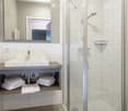 Modern bathroom with a sink, mirror, and a glass-enclosed shower area. The sink has a wooden shelf below holding neatly folded towels.
