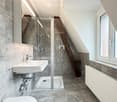 Modern bathroom with a white sink, chrome faucet, glass-enclosed shower, and radiator. The walls are tiled in grey marble, and the floor is made of dark stone tiles.
