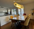 Modern kitchen and dining area with white cabinets, a wooden dining table, yellow chairs, and a pendant light fixture. The room features large windows with a view of the outside.