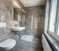 Modern bathroom with marble tiles, a toilet, a sink with a faucet, a shower stall, a mirror, and a radiator near the window.