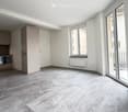 Open-plan living and kitchen area with grey tiled flooring, white walls, fitted kitchen cabinets, sink, and oven. Large window and sliding glass door leading to a balcony.