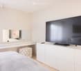 Modern bedroom with a large flat-screen TV on a white cabinet, a neatly made bed with striped bedding, a white dresser with a mirror, and a patterned stool.