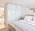 Modern bedroom with a large bed, nightstand, lamp, and sliding wardrobe. The room features light-colored walls, wooden flooring, and a view into an adjacent dining area with chairs and a table.
