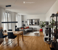 Modern living and dining area with wooden flooring, large windows, grey sectional sofa, dining table with chairs, cat tree, decorative shelf, and a vase with flowers.