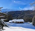 A snow-covered wooden house with a stone base, surrounded by snow-covered trees and mountains in the background.