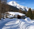 A large, snow-covered house with a stone facade and wooden elements, situated in a snowy mountain landscape with trees and a clear blue sky.