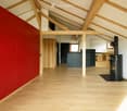 Open-plan attic space with exposed wooden beams, polished wooden flooring, and a red accent wall. Features a wooden door, a wooden table with chairs, a wooden cabinet, a wooden shelf, and a wood-burning stove.