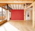 Bright, open room with wooden floors, exposed wooden beams, large windows offering a scenic view, and a red accent wall.