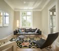 Spacious living room with large windows, hardwood floors, black leather sofa, patterned area rug, and decorative wall art