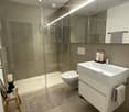 Modern bathroom with a glass-enclosed shower, white sink, toilet, and built-in vanity with mirrored cabinetry. Includes a small wooden stool with a candle and a beige bath mat.