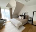 Modern bedroom with a large bed, grey armchair, side table, mirror, and decorative picture frames. The room features wooden flooring, a white rug, and large windows with city views.