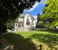 A large, elegant two-story house with white exterior and multiple windows, surrounded by a well-maintained lawn and trees. Features include a front staircase, a small porch area, and outdoor seating.