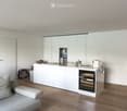 Modern, minimalist kitchen and living area with white cabinets, a central island, a wine cooler, and a grey sofa.