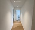A modern, minimalist hallway with light wooden flooring, white walls, and recessed lighting. An open door at the end leads to another room.