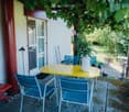 Outdoor seating area with a yellow table, blue chairs, and a red bench, surrounded by greenery and a covered entrance.