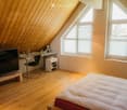 Bright attic bedroom with wooden floors and slanted wooden ceilings. Features a bed, desk with chair, TV on a stand, and a small shelf with books and decorative items.