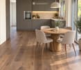 Modern dining area with a round wooden table surrounded by six upholstered chairs, pendant light fixture, grey cabinets, and a built-in oven. The space features light wooden flooring and large windows with a view of greenery.