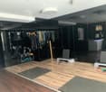 Modern fitness studio with wooden flooring, large mirrors, clothing rack, exercise bench, yoga mats, and weights.