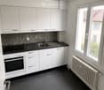 Modern kitchen with white cabinets, black countertop, stainless steel sink, and built-in oven. Dark tiled floor and radiator visible.
